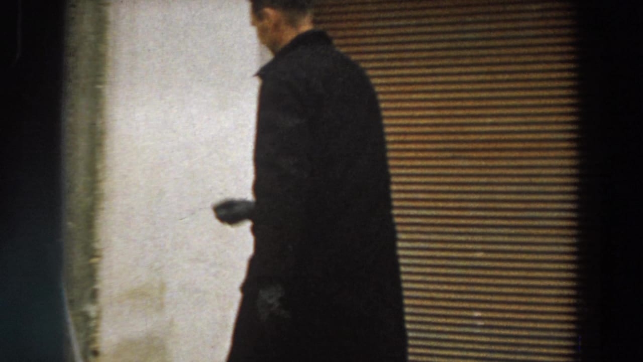 In 1958 Italy footage, a man in a dark overcoat passes a closed corrugated shutter along a rough exterior wall.