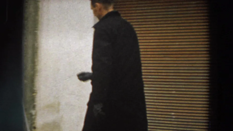 In 1958 Italy footage, a man in a dark overcoat passes a closed corrugated shutter along a rough exterior wall.