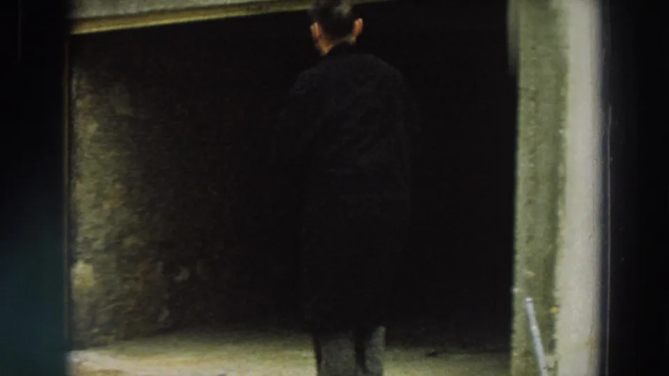 The man approaches or looks into a dark entrance beside a shuttered industrial-looking exterior in Italy.