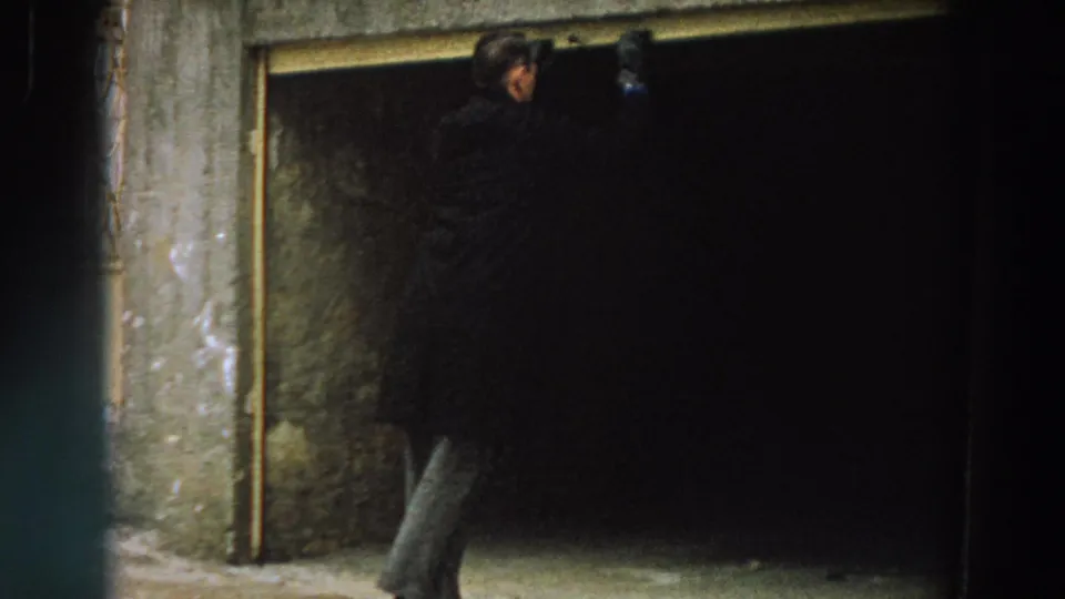 A man in a dark coat crosses in front of a shadowed opening, with worn exterior walls and pavement visible.
