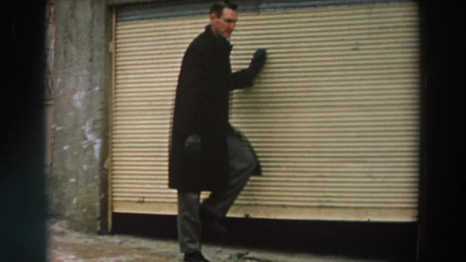 The man faces the camera while raising a hand near a closed corrugated shutter, with his overcoat and light trousers visible.