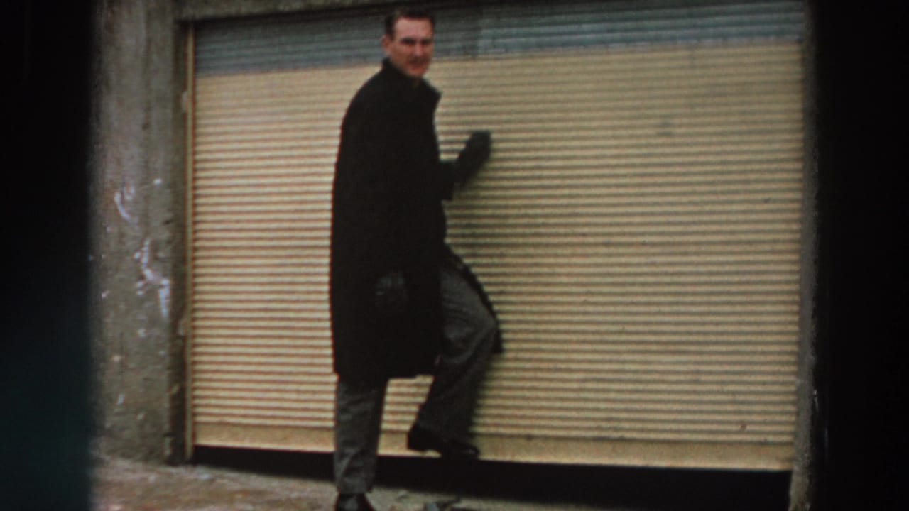 In 1958 Italy home-movie footage, a man in a dark overcoat and gloves poses near a closed metal shop shutter.