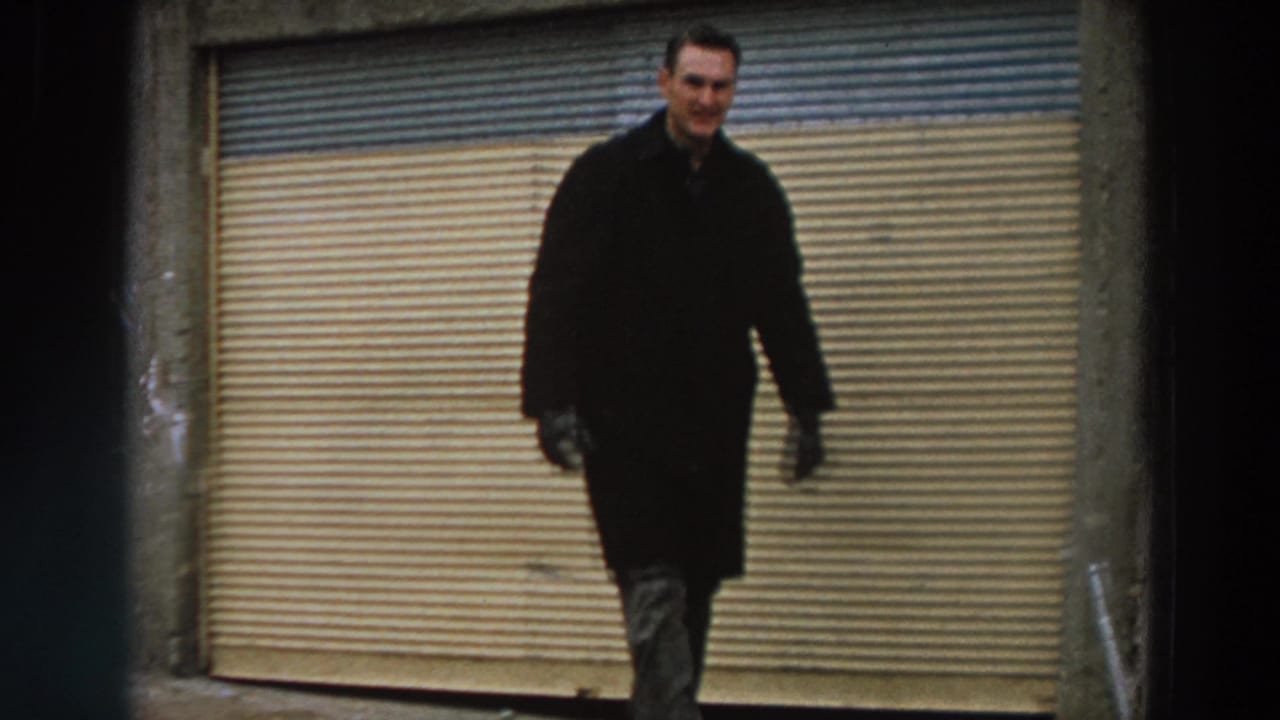 A smiling adult man in a dark coat walks toward the camera before a closed shutter in archival 1958 Italy footage.