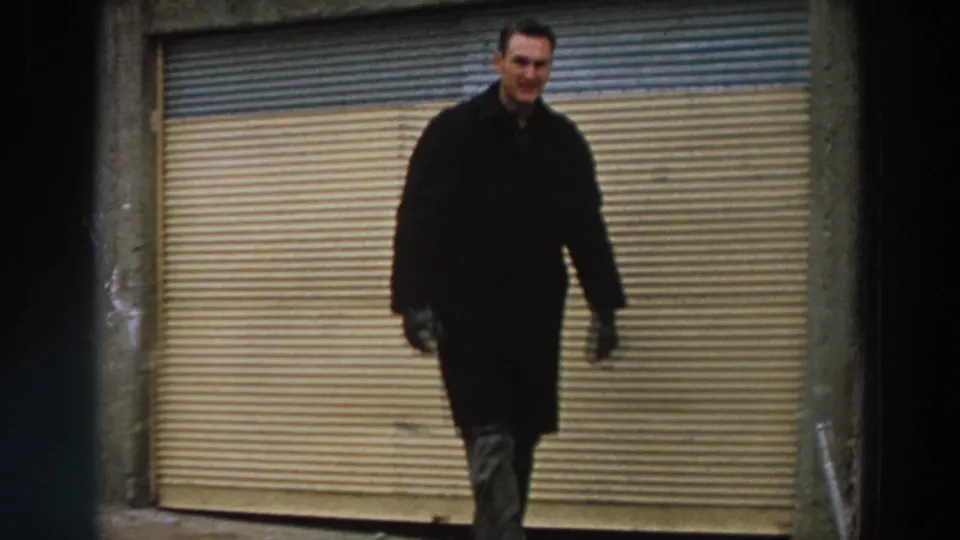 A smiling adult man in a dark coat walks toward the camera before a closed shutter in archival 1958 Italy footage.