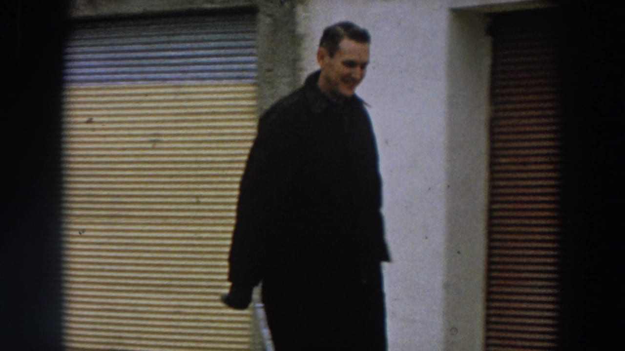 The man appears closer to the camera, framed against a pale wall and shutter, with soft 8mm grain and subdued color.