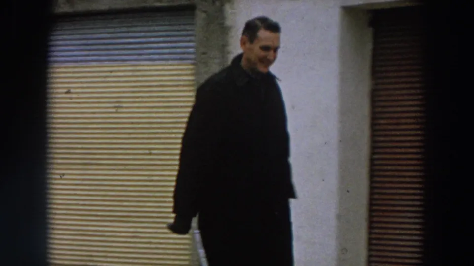 The man appears closer to the camera, framed against a pale wall and shutter, with soft 8mm grain and subdued color.