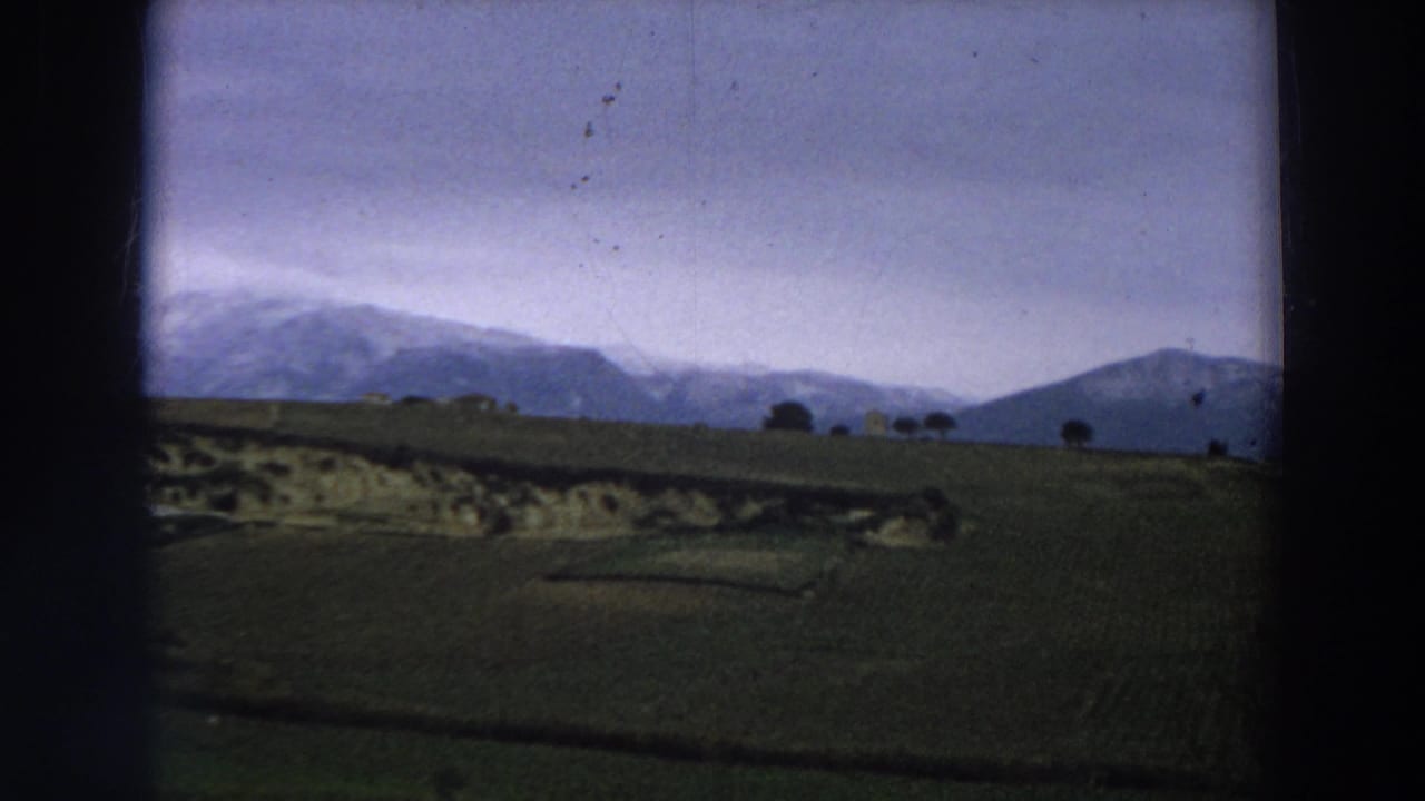 A rural Italian landscape shows green rolling fields, scattered trees, and distant mountains beneath a cloudy sky.