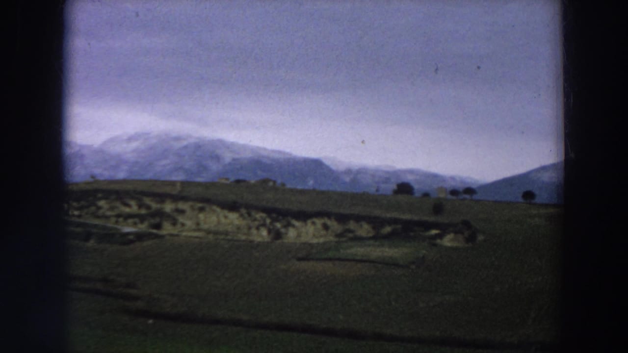 1958 Italy 8mm view of snow-covered mountains above green rural fields beneath a cloudy sky, with dark rounded film-frame edges.