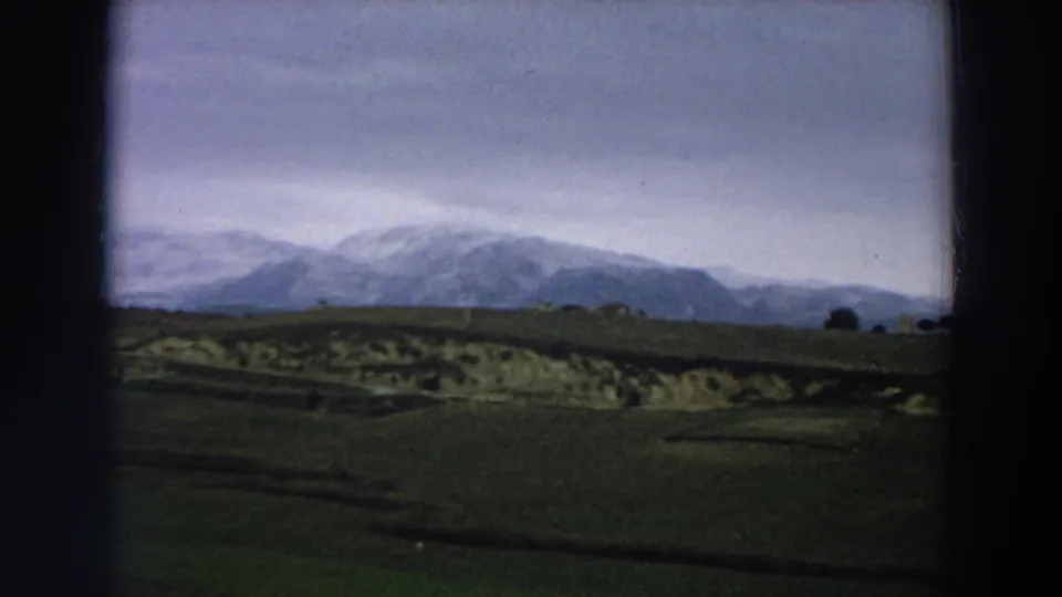 1958 Italy countryside view with rolling green fields, pale earth patches, scattered trees, and snow-covered mountains in the distance.