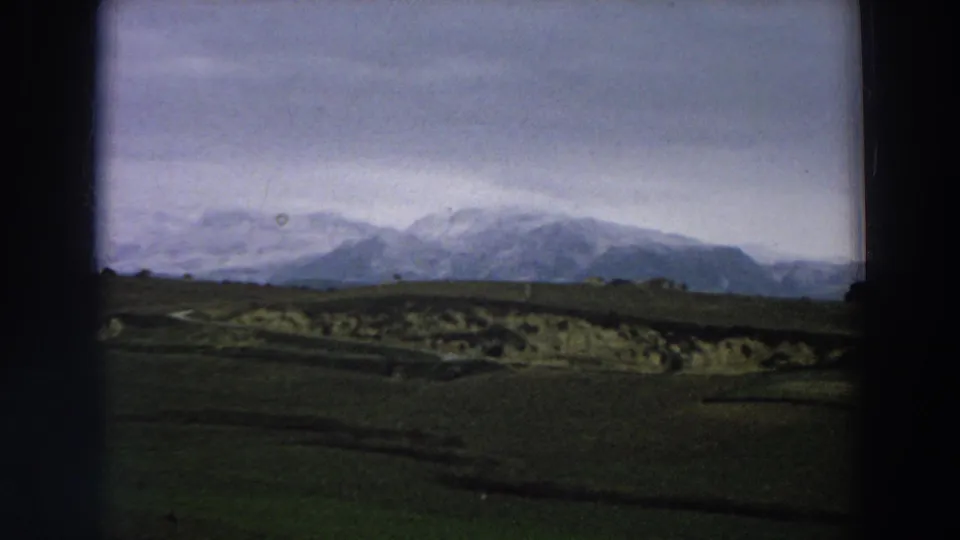 1958 Italy 8mm frame showing clouded mountain ridges, green cultivated-looking fields, pale exposed ground, and dark film borders.