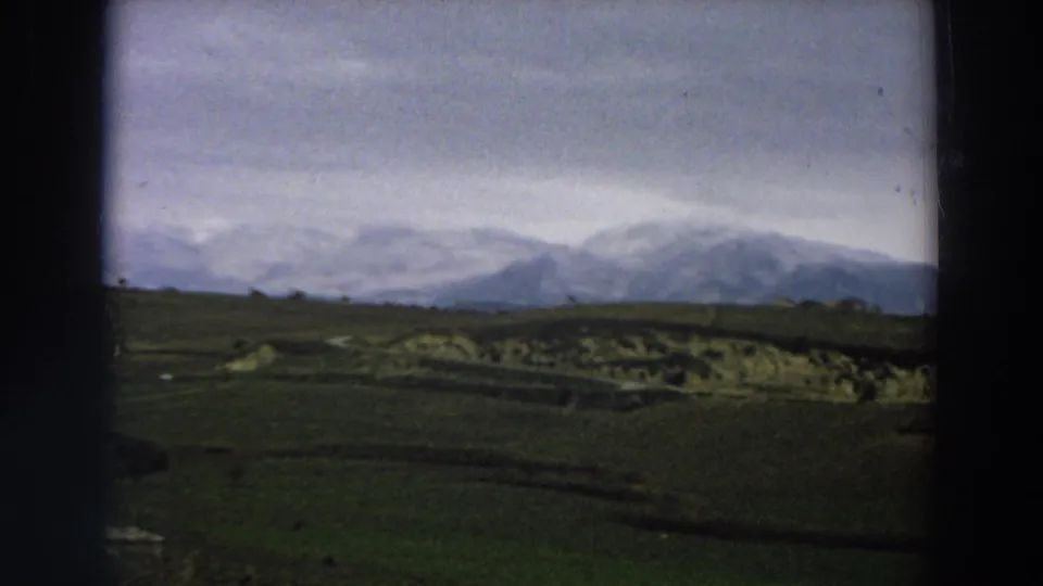1958 Italy landscape frame with snow-dusted mountains, broad green fields, low tree silhouettes, and visible 8mm film-frame borders.