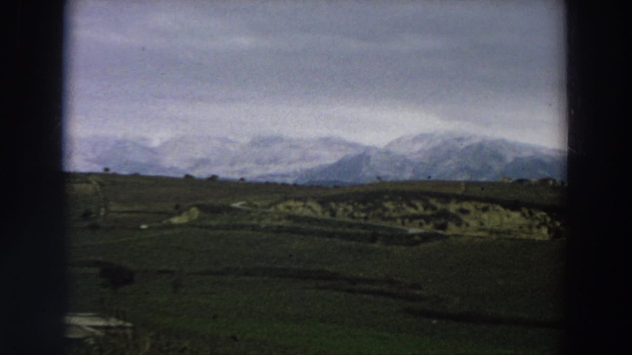 1958 Italy 8mm frame showing green fields, a small water area, distant structures, and snow-covered mountains under cloud.