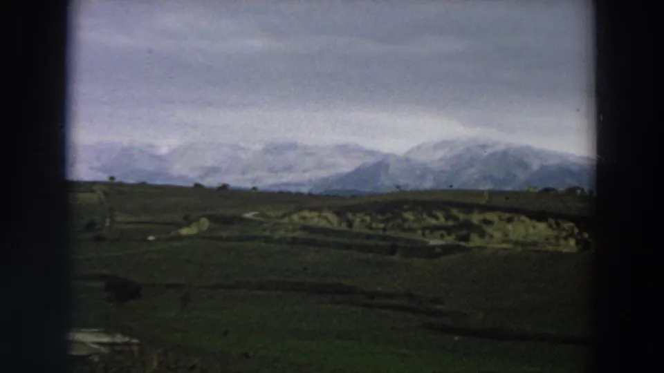 1958 Italy 8mm frame showing green fields, a small water area, distant structures, and snow-covered mountains under cloud.