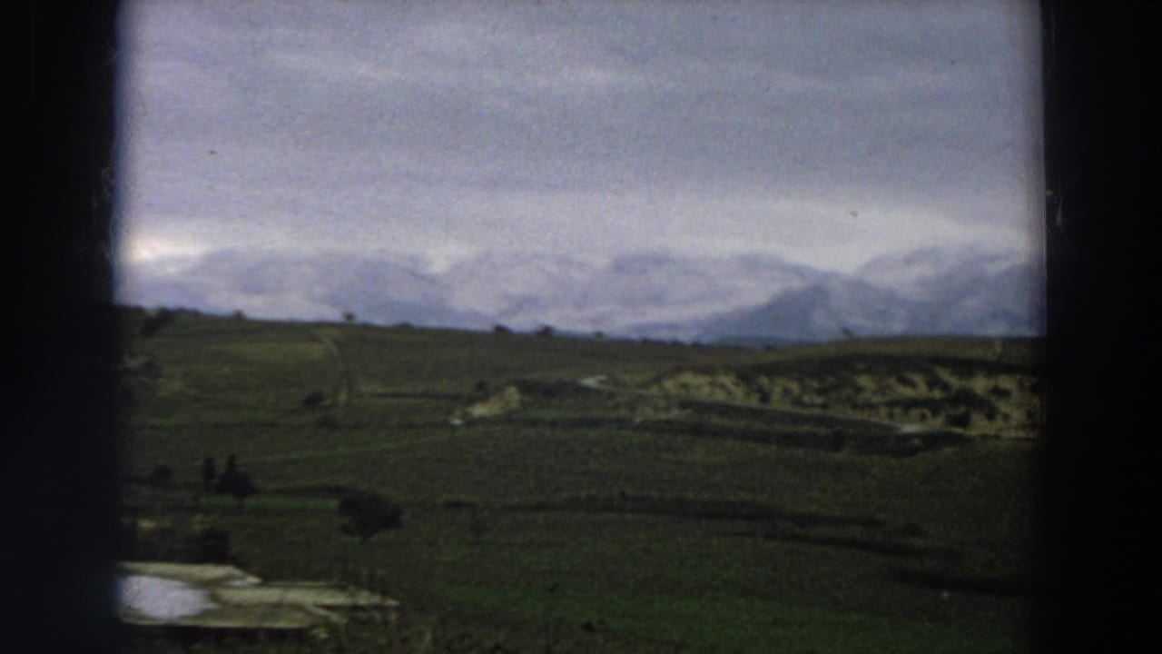 1958 Italy home-movie view of open fields, a reflective pond or lake edge, distant figures, and snowy mountains.
