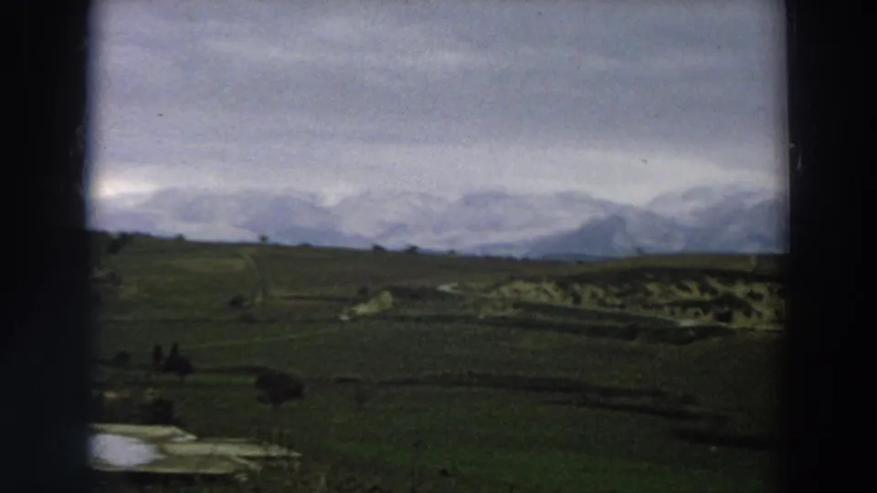 1958 Italy home-movie view of open fields, a reflective pond or lake edge, distant figures, and snowy mountains.