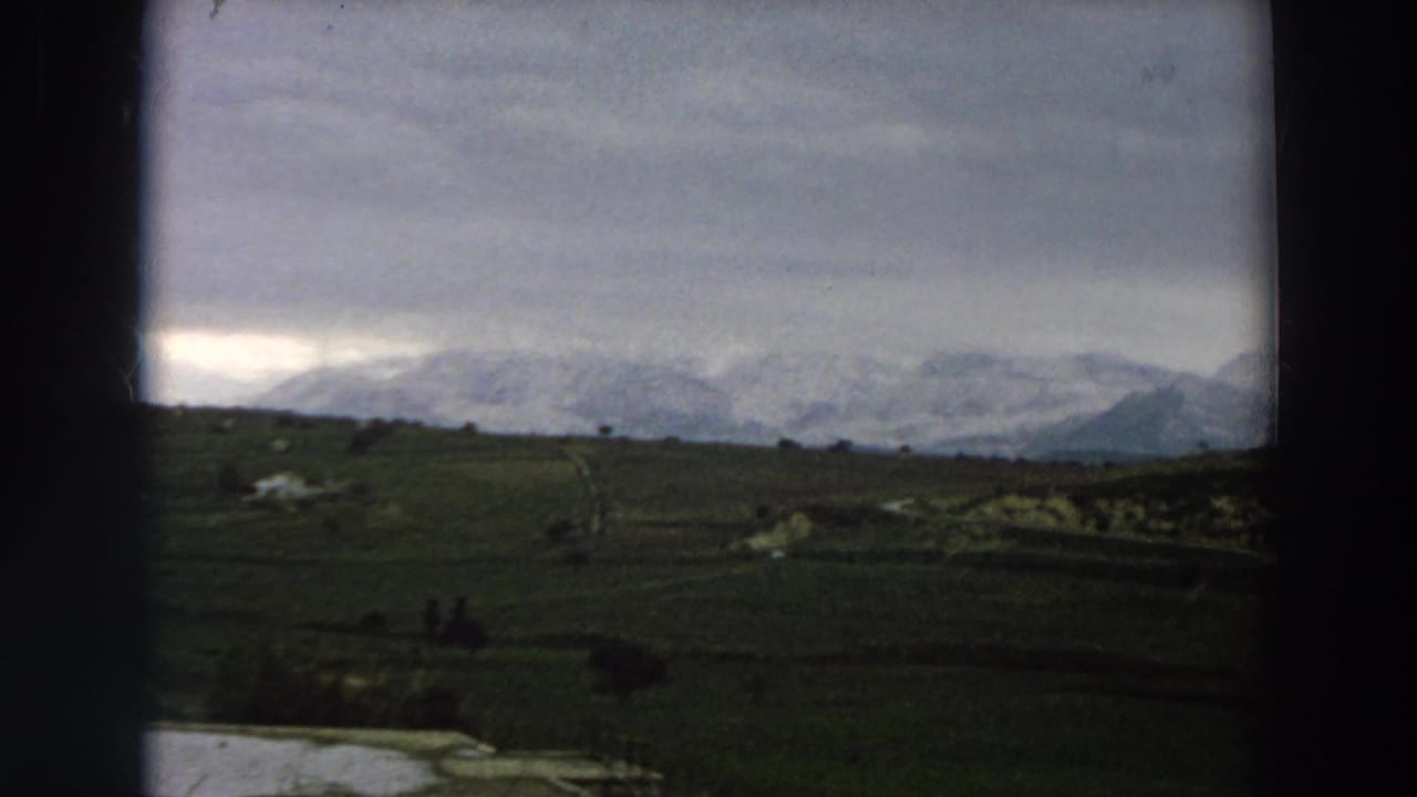 1958 Italy 8mm frame with dark meadow foreground, pale water at lower left, and snow-covered mountains on the horizon.