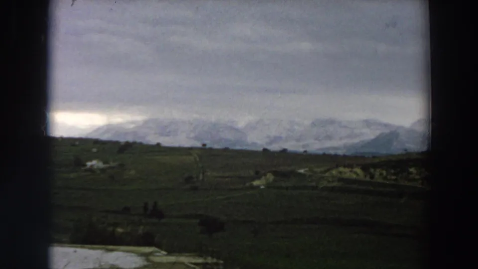 1958 Italy 8mm frame with dark meadow foreground, pale water at lower left, and snow-covered mountains on the horizon.