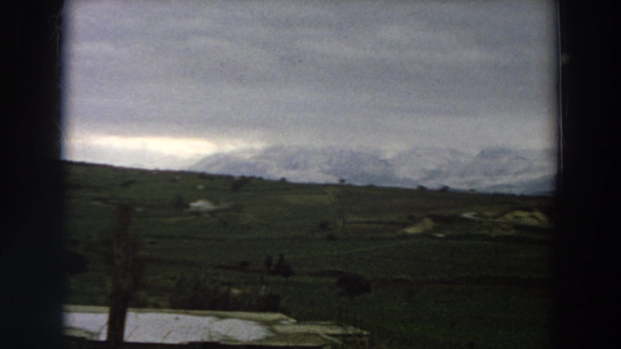 1958 Italy landscape frame showing fields, a small body of water, faint figures, and snowy mountains through soft 8mm color.