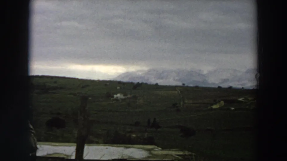 1958 Italy home-movie frame showing green farmland, fence posts, cloudy sky, and distant mountains.