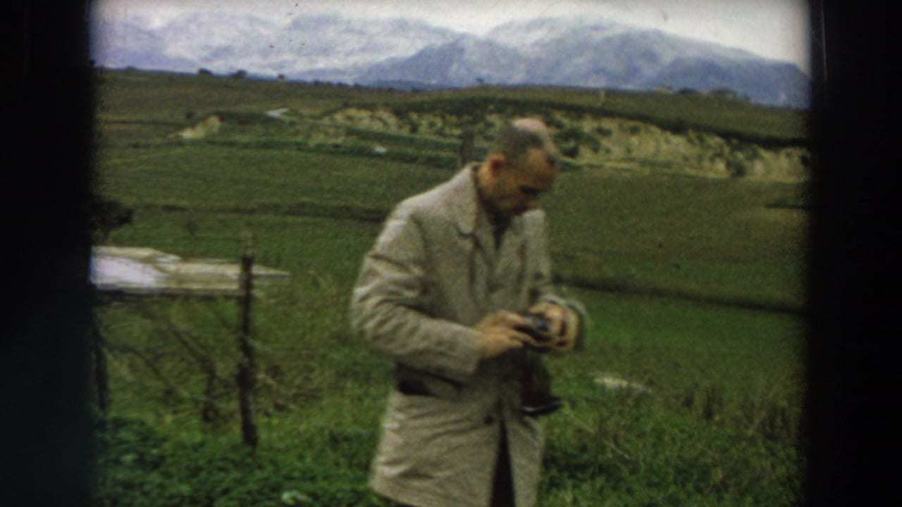 1958 Italy frame of an adult man in a light coat handling camera equipment in a rural field with mountains behind him.