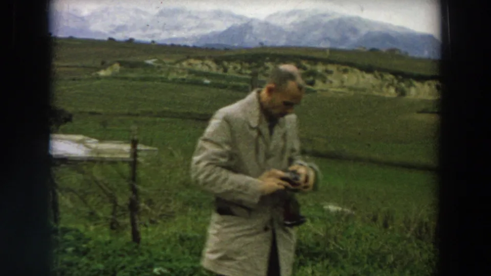 1958 Italy frame of an adult man in a light coat handling camera equipment in a rural field with mountains behind him.