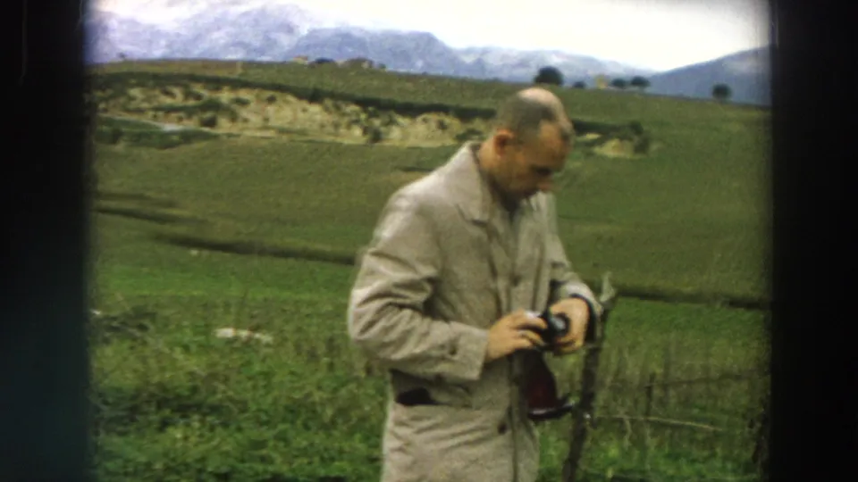 1958 Italy home-movie frame showing a man focused on his camera near cultivated fields and low hills.