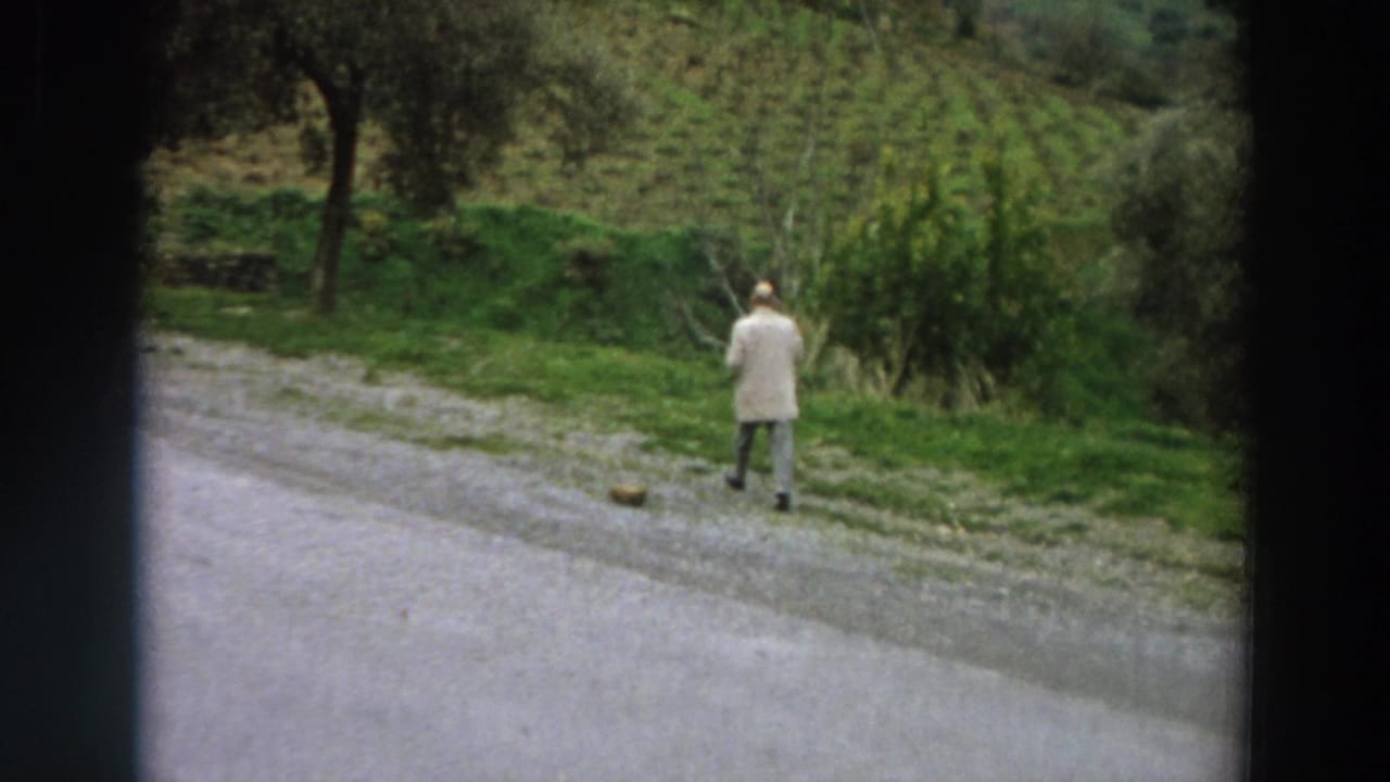 1958 Italy home-movie frame showing a man in a light jacket walking away beside a gravel road and green hillside plants.
