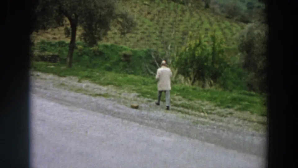 1958 Italy home-movie frame showing a man in a light jacket walking away beside a gravel road and green hillside plants.