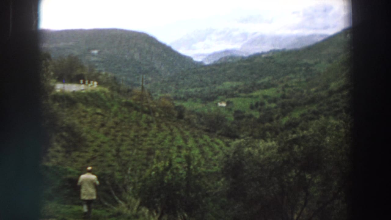 1958 Italy home-movie frame showing a distant man near a hillside crop field with mountains and clouds beyond.