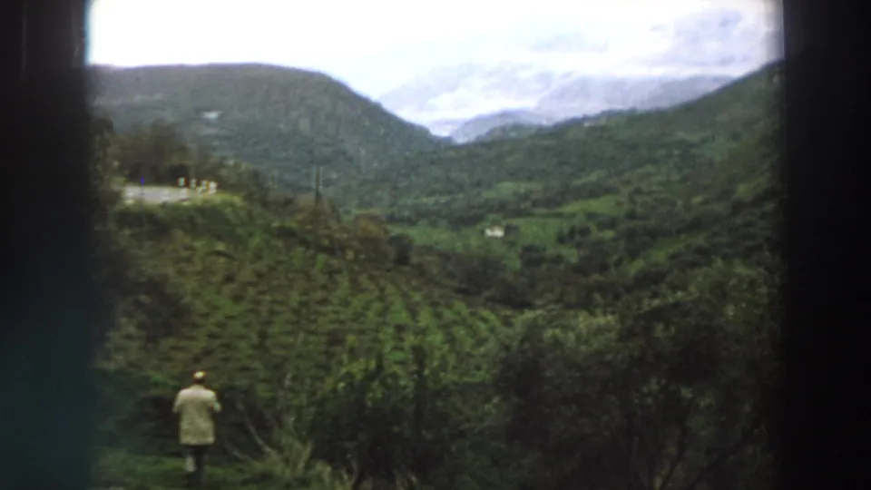 1958 Italy home-movie frame showing a distant man near a hillside crop field with mountains and clouds beyond.