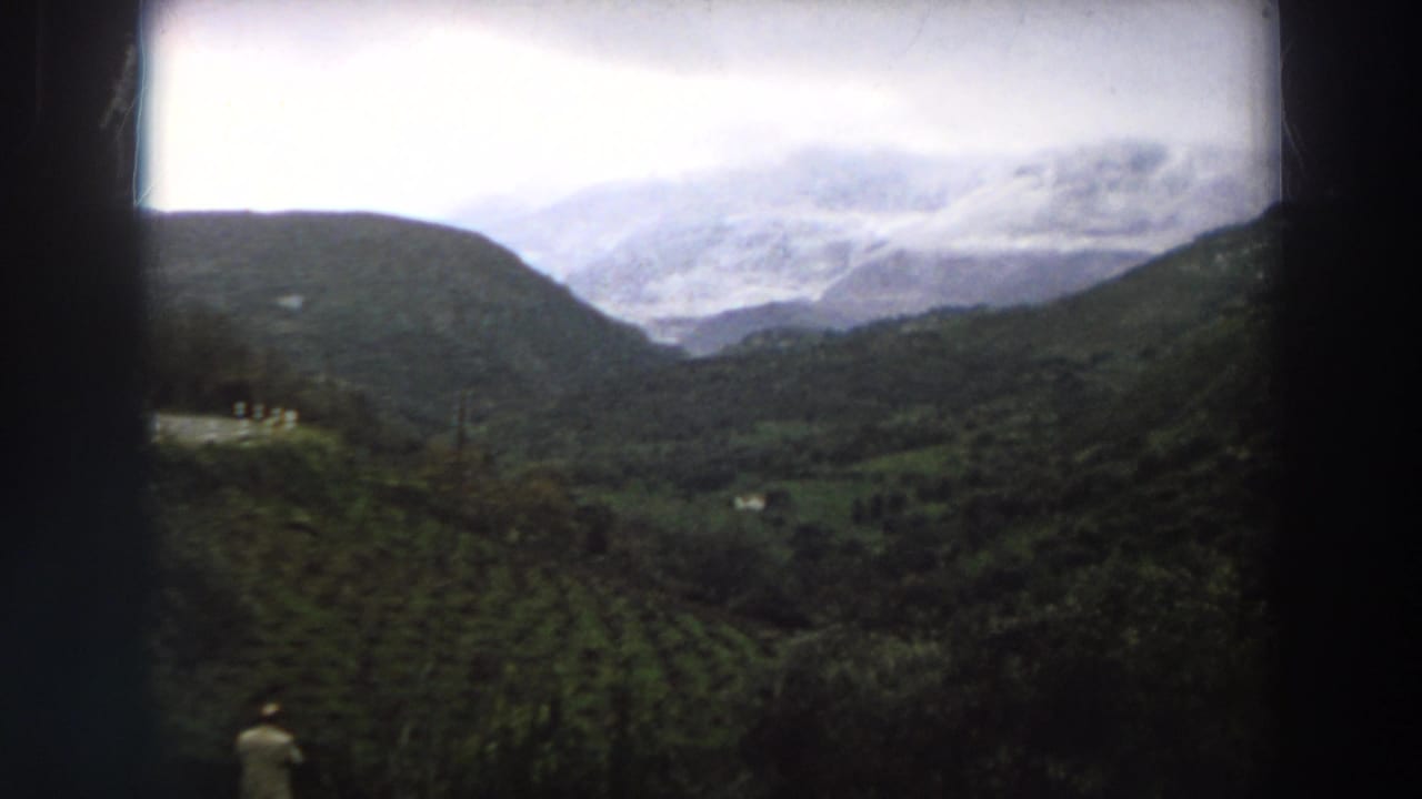 1958 Italy home-movie frame presenting a wide mountain valley with green fields, trees, and overcast cloud cover.