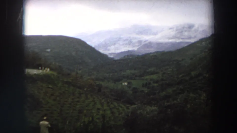 1958 Italy home-movie frame presenting a wide mountain valley with green fields, trees, and overcast cloud cover.