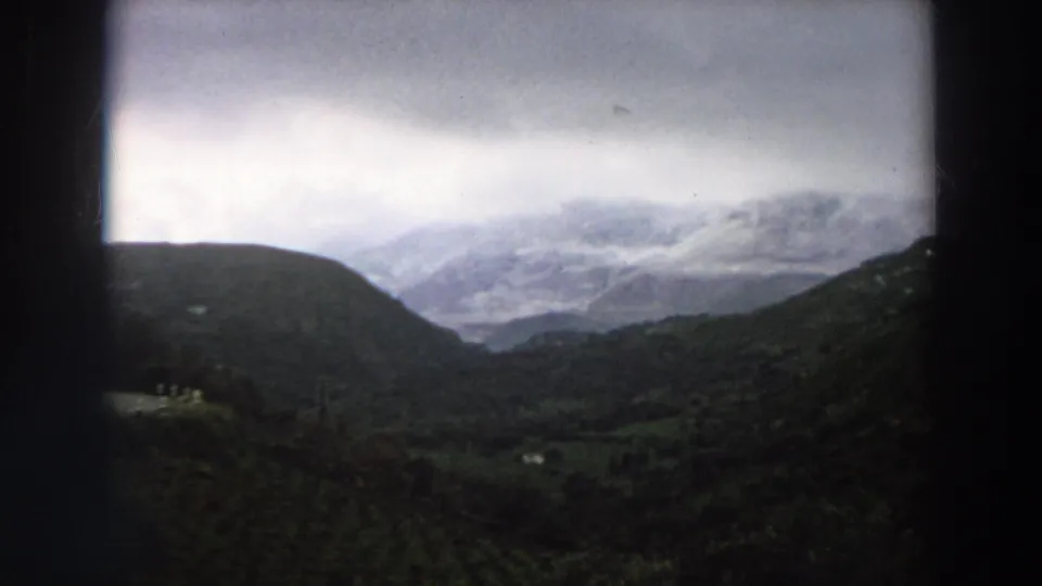 1958 Italy home-movie frame showing a cloudy mountain vista with dark film borders and green hills in the foreground.