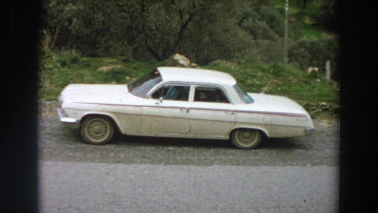 A white mid-century sedan is parked at a roadside in Italy, framed by trees, grass, and visible 8mm film edges.