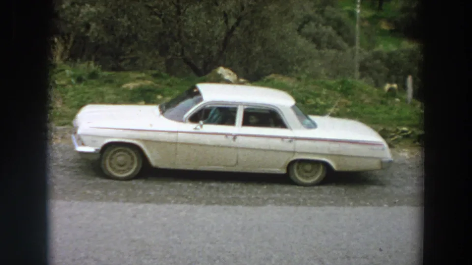 A white mid-century sedan is parked at a roadside in Italy, framed by trees, grass, and visible 8mm film edges.