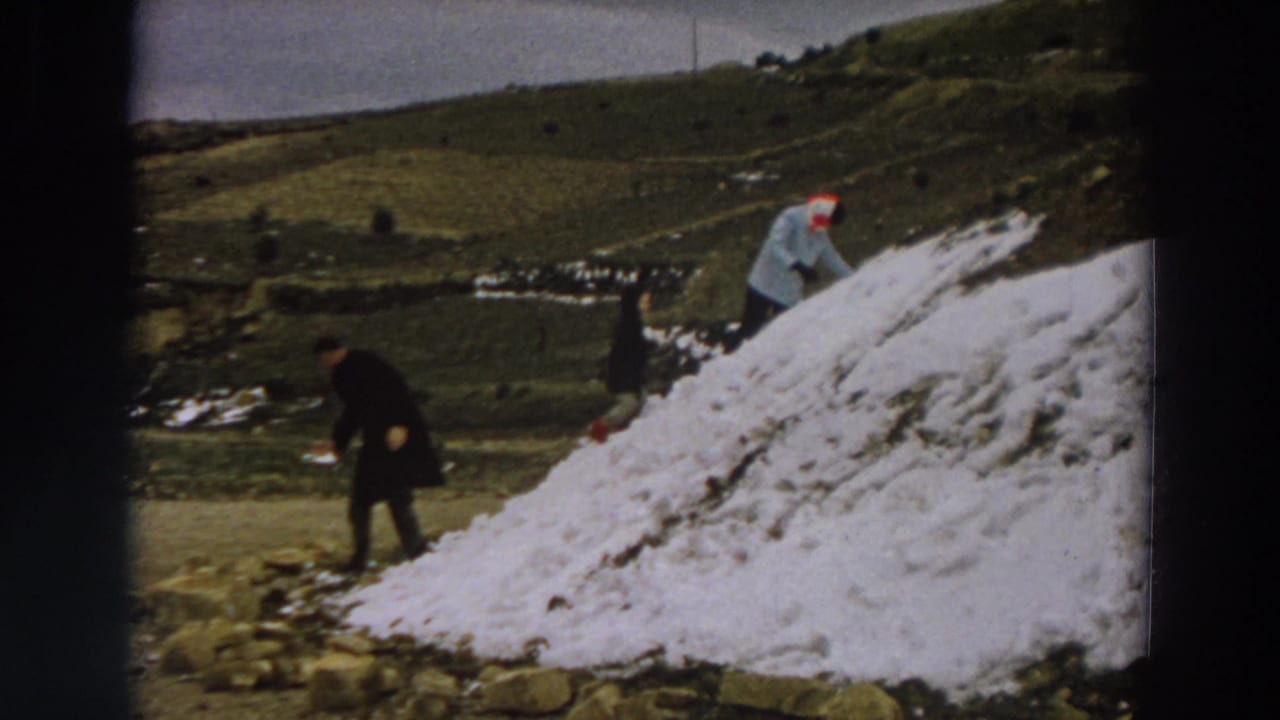 Two people work around a large snow pile on a grassy hillside.