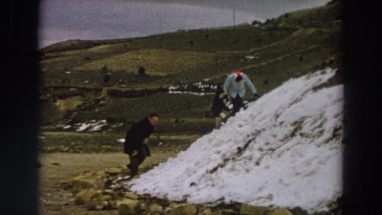 1958 Italy 8mm still showing figures around a snow pile with rolling green hills and scattered snow behind them.