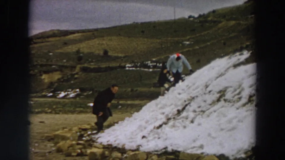 1958 Italy 8mm still showing figures around a snow pile with rolling green hills and scattered snow behind them.