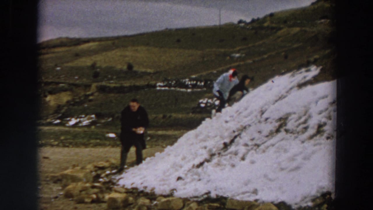 1958 Italy home-movie frame with a dark-coated figure near the camera and another person crouched on the snowy mound.
