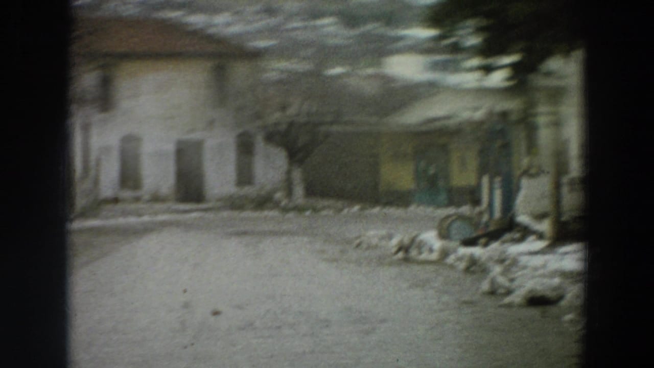 1958 Italy home-movie frame showing a blurred approach to pale street buildings, a yellow facade, green doorway, bare trees, and patches of snow.