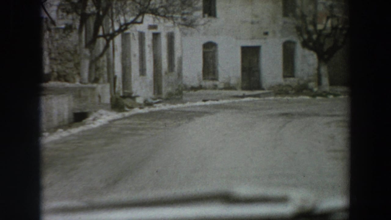 1958 Italy street view with pale stucco facades, dark doorways, bare trees, a curving road, and snow along the edges.