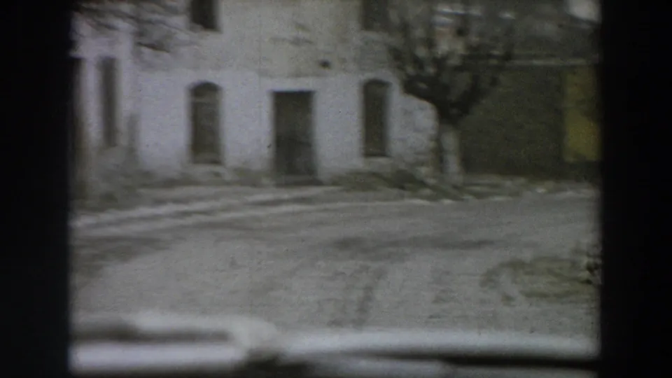 1958 Italy frame showing a soft, moving street-corner view of pale building fronts, shuttered openings, a bare tree, and winter ground patches.