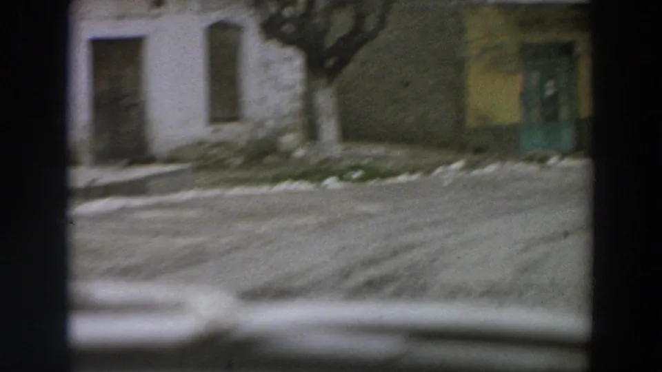 1958 Italy home-movie frame with a white wall, dark doorway, bare tree trunk, yellow building surface, green door, and road edge in soft motion.