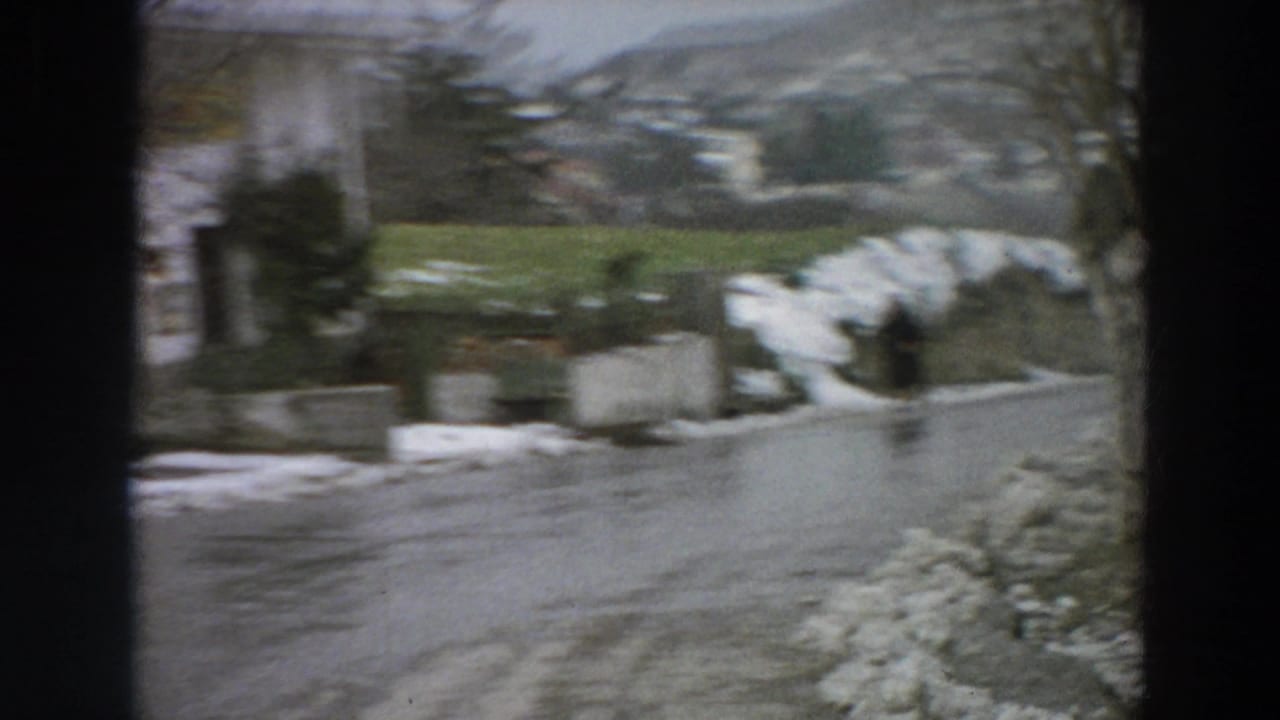 1958 Italian 8mm frame from a moving car on a wet icy village road with snowbanks, walls, trees, and hillside homes beyond.