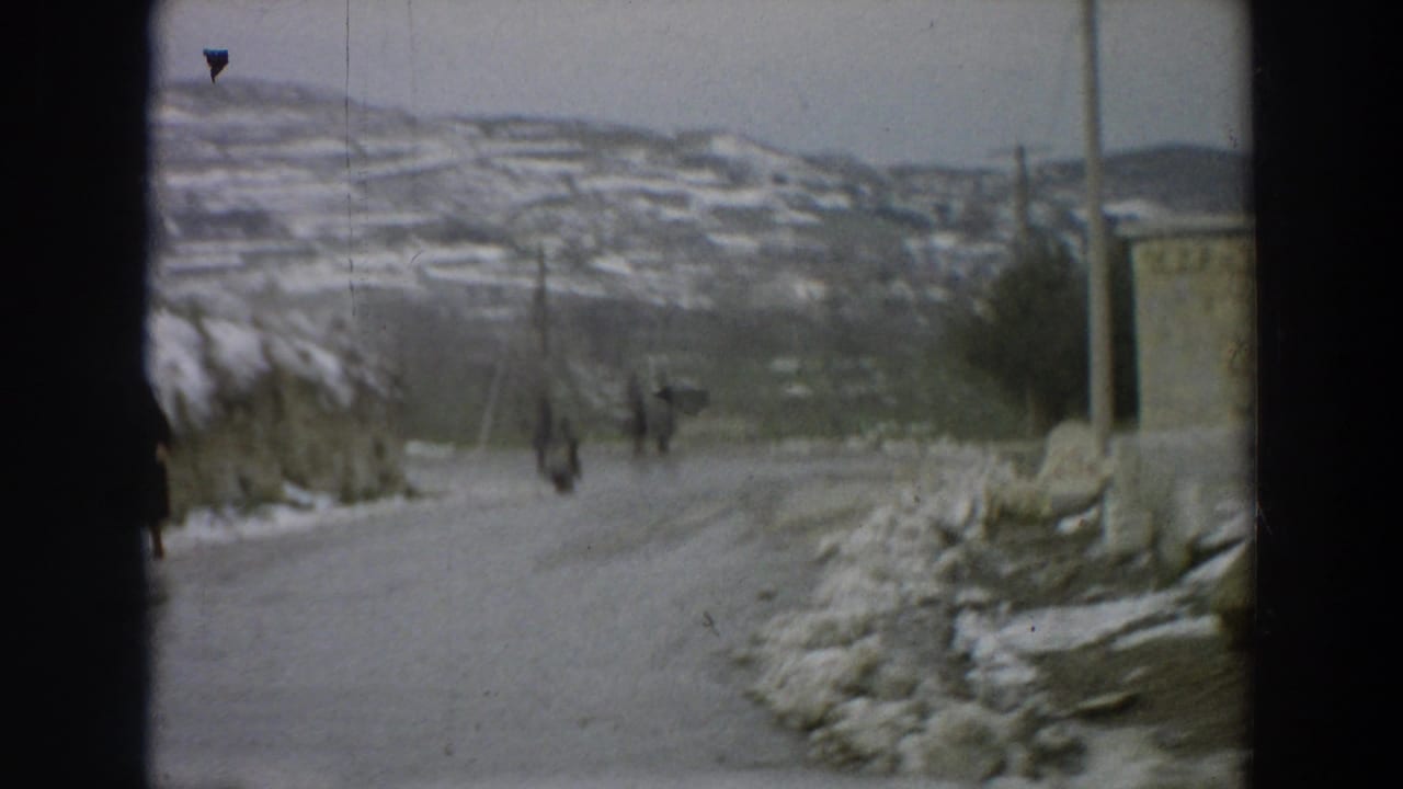 Snowy road with pedestrians in the distance, utility poles, hillside houses, and a roadside building.