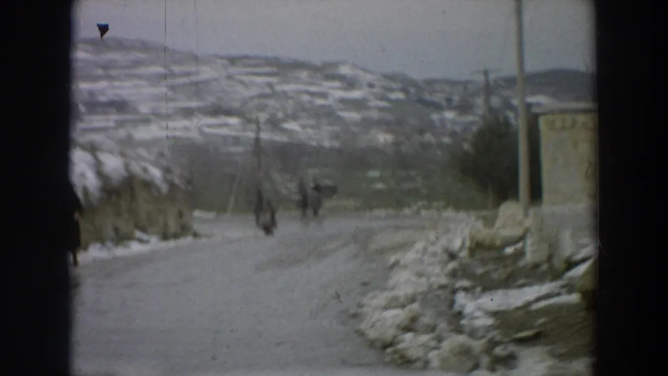 1958 Italian village road scene with distant pedestrians walking on an icy route below snow-dusted hillside houses.