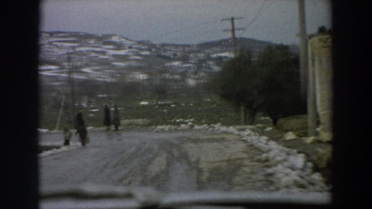 1958 Italian 8mm frame showing pedestrians on a curving winter village road with snowbanks, poles, trees, and hillside dwellings.