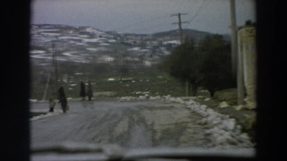 1958 Italian 8mm frame showing pedestrians on a curving winter village road with snowbanks, poles, trees, and hillside dwellings.
