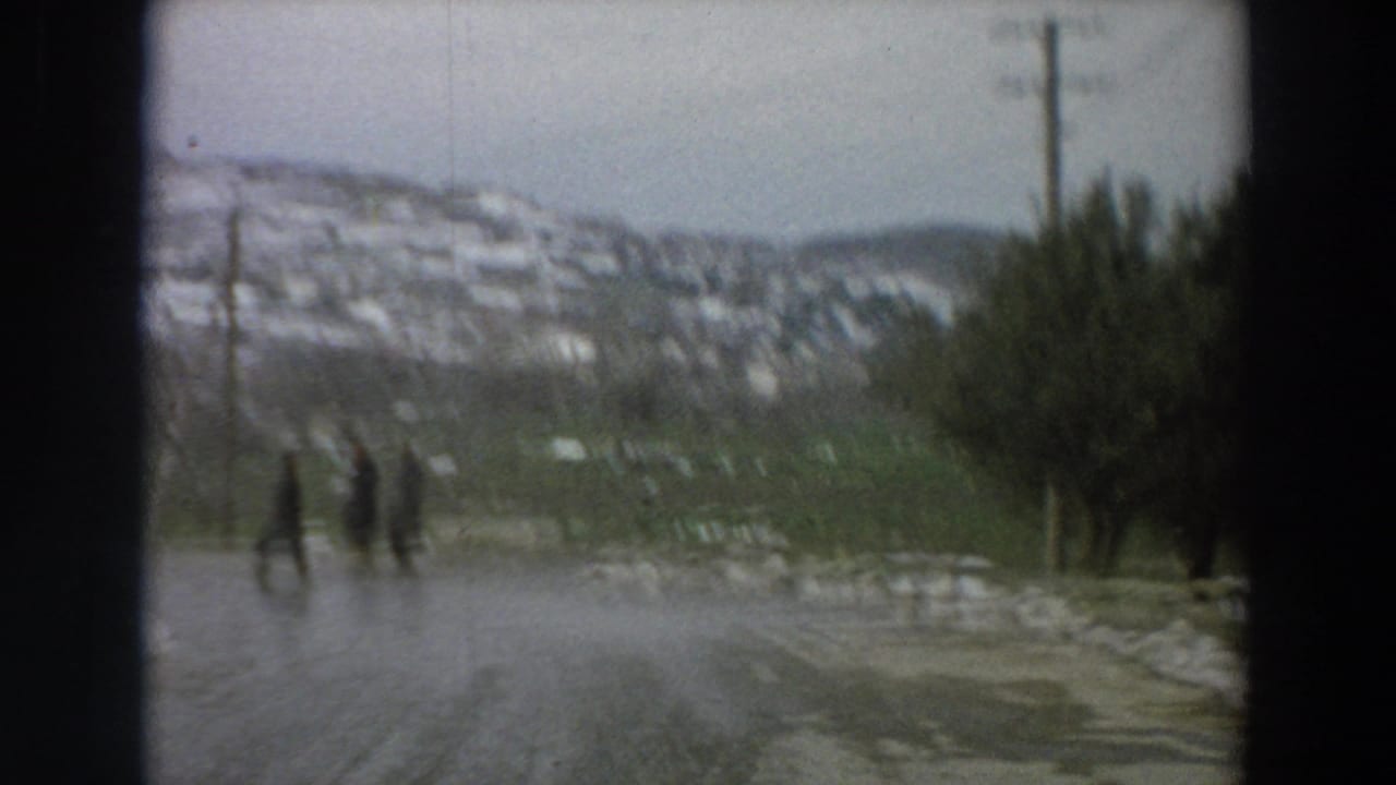 Soft 1958 Italian village road frame with distant pedestrians, wet icy pavement, snowy hillside houses, and winter trees.