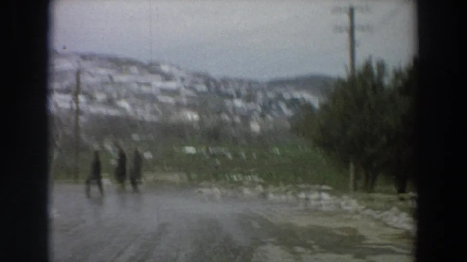 Soft 1958 Italian village road frame with distant pedestrians, wet icy pavement, snowy hillside houses, and winter trees.